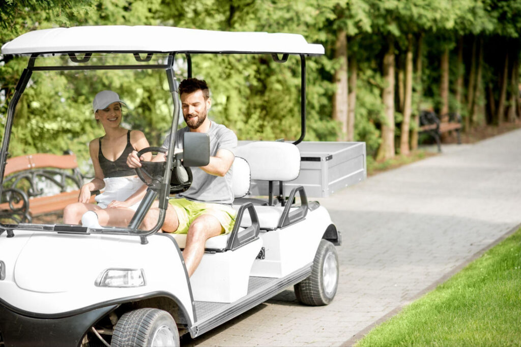 Young couple driving a white electric golf cart rental for two in Bradenton, Florida, enjoying a sunny day ride along park paths with trees and benches in the background.Picture