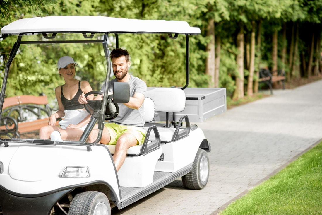 Young couple driving a white electric golf cart rental for two in Bradenton, Florida, enjoying a sunny day ride along park paths with trees and benches in the background.Picture