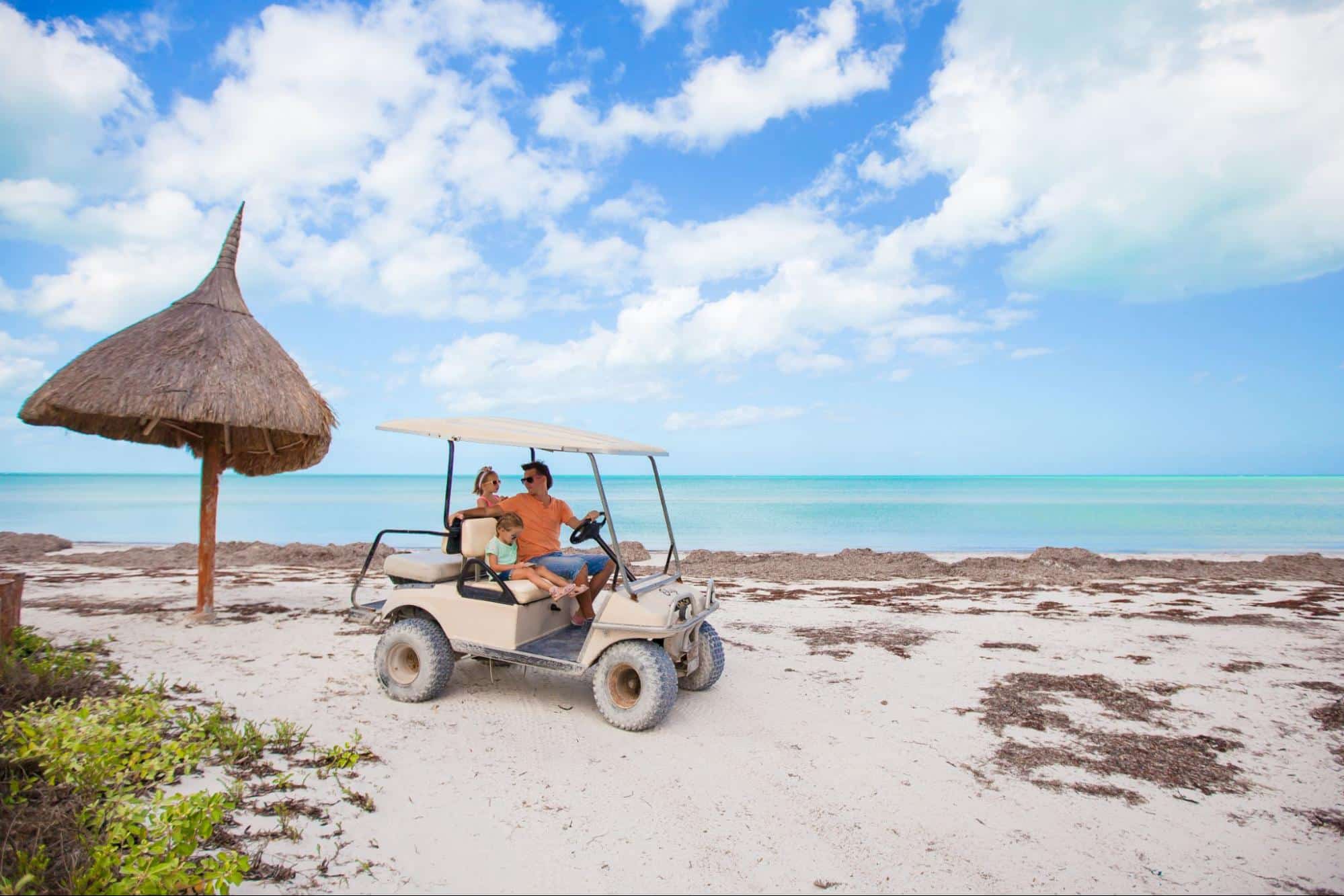 Family with young daughter exploring a tropical beach by golf cart with turquoise water and white sand.